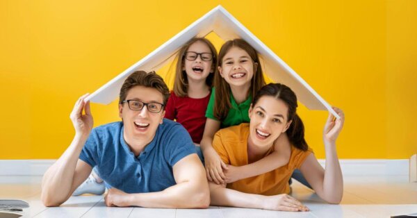 Family opportunity mortgage: Four people holding a model house roof over their heads in a room with a yellow wall.