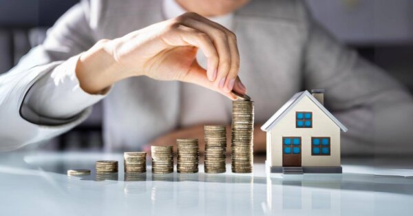 Person stacking coins beside a model house, symbolizing real estate investment or savings, with a focus on luxury real estate funds as a profitable opportunity.