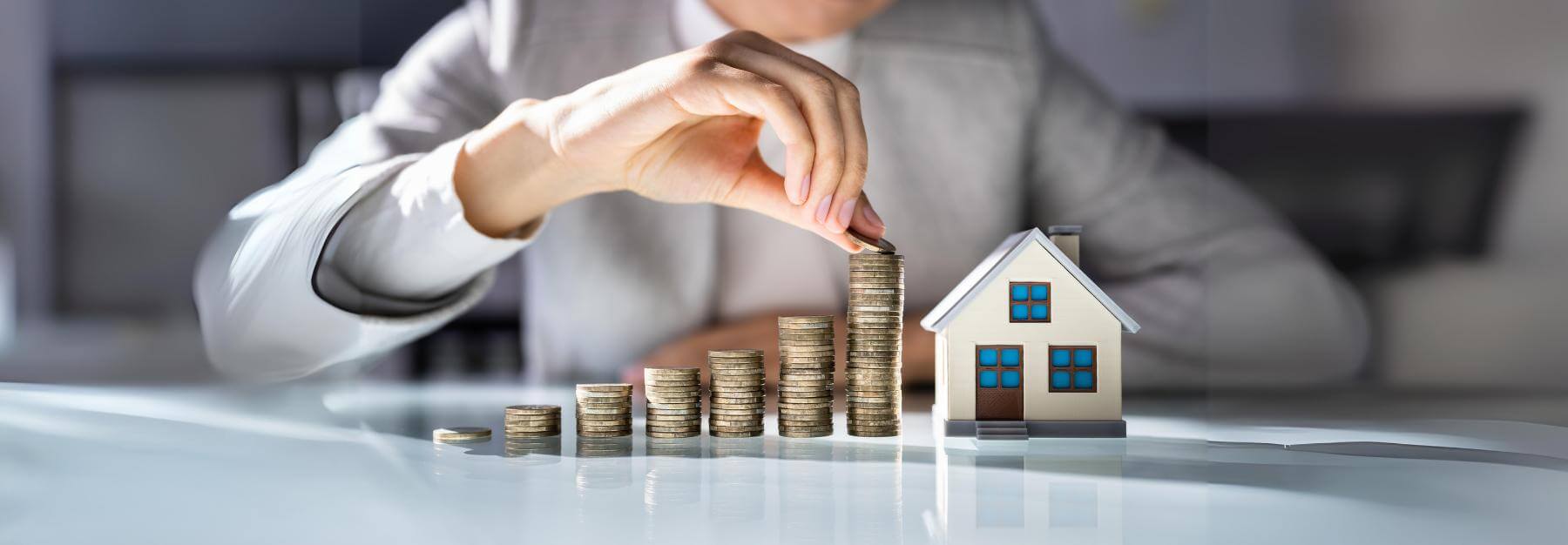 Person stacking coins beside a model house, symbolizing real estate investment or savings, with a focus on luxury real estate funds as a profitable opportunity.