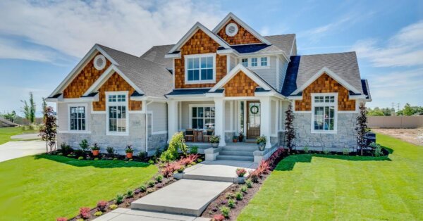 New two-story suburban house with a stone and wooden façade, a beautifully landscaped lawn, a shed with porch, and a clear blue sky.