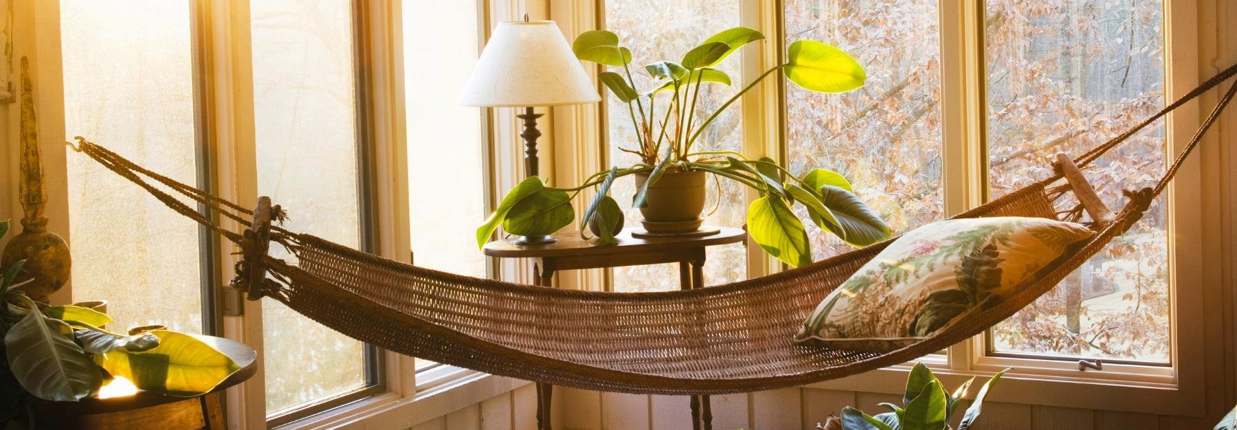 A cozy hammock with a pillow beside a sunroom windows with plants and lamp, overlooking autumn trees.